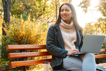 Sticker - Smiling young asian woman wearing coat sitting on a bench