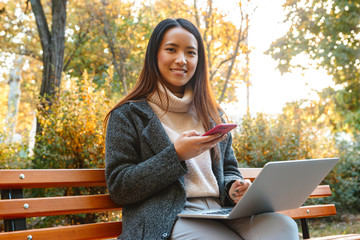 Sticker - Smiling young asian woman wearing coat sitting on a bench