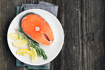 Fresh salmon steak on light plate with lemon and rosemary on wooden background. Top view.