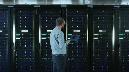 Poster - Bearded IT Specialist in Glasses is Working on Laptop in Data Center while Standing Before Server Rack. Running Diagnostics or Doing Maintenance Work. Camera Zooms Out.