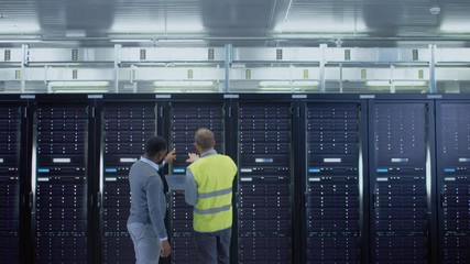 Aufkleber - Bearded IT Specialist in Glasses and High Visibility Vest with a Laptop Computer and Black Technician Colleague Talking in Data Center while Standing Next to Server Racks. Running Diagnostics. 