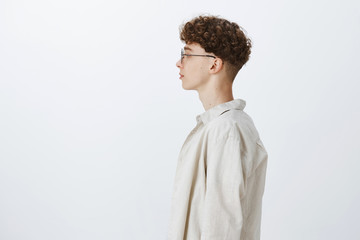 Profile shot of stylish handsome young hipster male freelancer with curly hairstyle in glasses looking left and standing casually over gray background in shirt, having no emotions as feeling calm