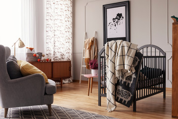 Blanket on bed next to armchair in grey baby's bedroom interior with poster and window. Real photo