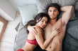 Handsome satisfied young man lying on bed with beautiful model. She embrace him and smile. Young woman is sleep. Guy hold hand behind head.
