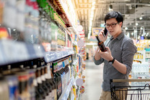 Asian Man Using Phone Shopping Beer In Supermarket. Male Shopper With Shopping Cart Choosing Beer Bottle In Grocery Store.