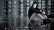 Reflection of mystery hoodie man in white mask hugging his knees sitting in the rain on rooftop of abandoned building. Bipolar disorder or Major depressive disorder. Depression concept