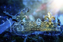 Mysterious And Magical Photo Of Silver King Crown And Sword Over The Stone Covered With Moss In The England Woods Or Field Landscape With Light Flare. Medieval Period Concept.