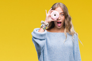 Wall Mural - Beautiful young blonde woman eating pink donut over isolated background scared in shock with a surprise face, afraid and excited with fear expression