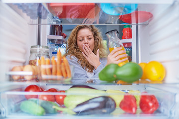 Wall Mural - Woman standing in front of fridge full of groceries and taking juice. Picture taken from inside of fridge.