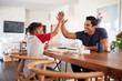 © Monkey Business - Hispanic father and son sitting opposite each other, high five over the dining room table, side view
