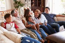 Three Generation Hispanic Family Sitting On The Sofa Watching TV, Grandmother Using Remote Control