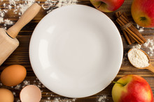 Empty White Plate And Ingredients For Baking Apple Pie On The Brown Wooden Background.Top View.Copy Space.