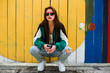 © Nabokov Alex - Portrait of a girl in clothes in the style of the 90s, sporty style, jacket, jeans, bananas, sunglasses. Lady on the background of wooden colored background, drinking morning coffee