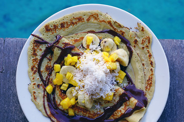 Wall Mural - A tropical breakfast crepe with pineapple, chocolate, and fresh coconut eaten by the blue sea