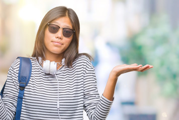 Poster - Young asian student woman wearing headphones and backpack over isolated background clueless and confused expression with arms and hands raised. Doubt concept.