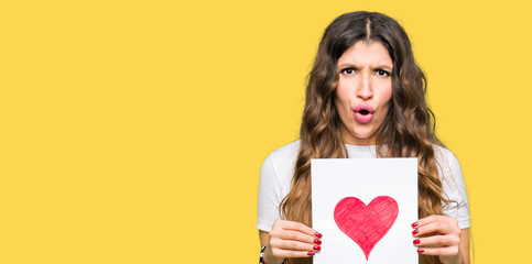 Sticker - Young adult woman holding card with red heart scared in shock with a surprise face, afraid and excited with fear expression