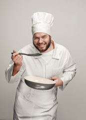 Poster - Handsome bearded guy in chef uniform smiling and looking at camera while standing on light gray background and trying food from frying pan