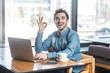 © khosrork - Portrait of handsome positive bearded young freelancer in blue jeans shirt are sitting in cafe and working on laptop with toothy smile and showing okay sign, looking at camera.