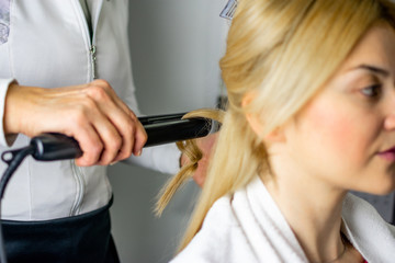 Wall Mural - Hairdresser holding strand of girls blonde hair with flat iron making beautiful curls