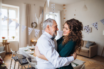 Senior man and young woman standing indoors in a room set for a party, hugging.