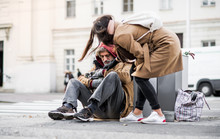 Young Woman Giving Money To Homeless Beggar Man Sitting In City.