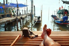 Part Of Tourist Legs In Relaxing Gesture And Camera Bag On Bamboo Litter With Traditional Fishing Boats Moored Over Seaside View And Sunrise Sky Background At Home Stay In Ecotourism Concept