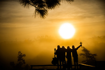 Wall Mural - Youth christian group holding cross and guitars with light sun rise background for worship god in morning, christian silhouette concept.