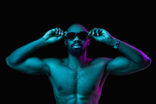 Close Up Portrait Of A Young Naked Happy Smiling African Man In Sunglasses At Studio. High Fashion Male Model In Colorful Bright Lights Posing On Black Background. Art Design Concept