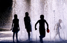 Silhouette Of Four Children Playing In A Water Fountain On A Hot Day, United States