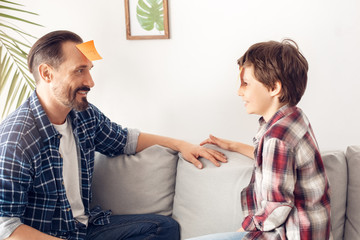 Father and little son at home sitting on sofa playing forehead detective guessing words cheerful side view