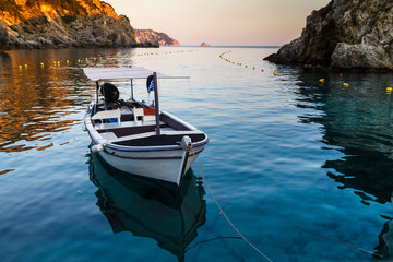 Wall Mural - The boat moored in the sea near the rocky coast, reflected in the water. Beautiful sunset light, rocks in the background. Holidays in Greece, Corfu island.