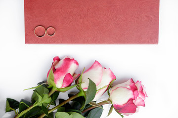 Red leather covered wedding book or album with two wedding rings and three roses lies on white background.