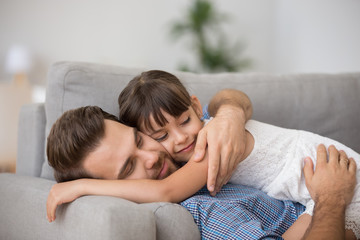 Canvas Print - Happy father hugging cute little girl lying together resting on comfortable sofa, smiling dad embracing kid daughter cuddling enjoying healthy daytime nap at home, daddy and child relaxing on couch