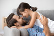 Cute little girl tenderly touching noses with happy dad lying on sofa, kid daughter and father having fun bonding feeling love connection enjoy relaxing together, daddy and child warm relationships