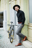 Fototapeta Łazienka - Confident and stylish. Full length of confident young bearded man adjusting eyewear and hat and looking at camera while standing near his bicycle outdoors