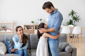 Happy father embracing caressing cute daughter, family spending time at home, parents with kid girl enjoy weekend together, cheerful mommy daddy and child laughing having fun playing in living room