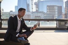Millennial Businessman Wearing Black Suit And White Shirt Sitting On The River Thames Embankment Using Smartphone, Close Up