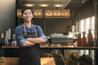 © Sirichai - Successful small business owner standing with crossed arms with employee in background preparing coffee. Male Owner Of Coffee Shop.