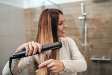 Beautiful Young Woman Using Hair Straightener And Smiling While Looking Into The Mirror In Bathroom. Focus On The Foreground, On The Hair Straightener.