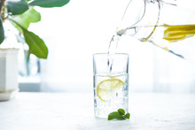 Pouring A Glass Of Water With Lemon, Ice And Mint On A White Table