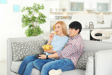 Wall Mural - Young couple with bowl of chips watching TV on sofa at home