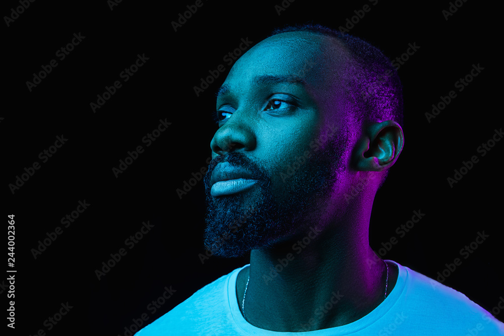 The retro wave or synth wave portrait of a young happy serious african man at studio. High Fashion male model in colorful bright neon lights posing on black background. Art design concept - obrazy, fototapety, plakaty 