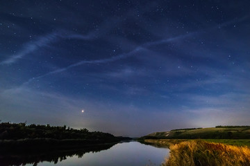 Wall Mural - Clouds in the night sky illuminated by the moon over a meadow and a river.