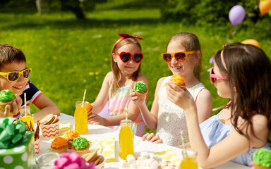 Poster - holidays, childhood and celebration concept - happy kids in sunglasses sitting at table on birthday party at summer garden and eating cupcakes