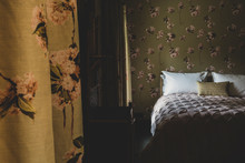 Interior View Of Bedroom With Curtains And Wallpaper With Floral Pattern, Pale Pink Quilt And White Pillows On Double Bed.