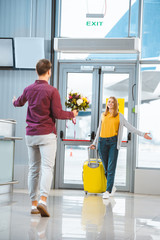 Wall Mural - back view of boyfriend with flowers meeting beautiful girlfriend with suitcase in airport