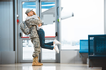 Wall Mural - Happy boyfriend in military uniform holding in arms girlfriend in airport