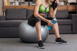 © LIGHTFIELD STUDIOS - cropped view of tired sportswoman sitting on fitness ball with towel and sport bottle at home
