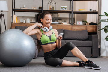 Wall Mural - smiling sportswoman in headphones sitting with fitness ball and using smartphone at home