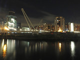 Fototapeta  - a cityscape view of the canal entrance to the clarence dock area of leeds with a pedestrian bridge crossing the water with reflections of lights and buildings against a night sky with clouds
